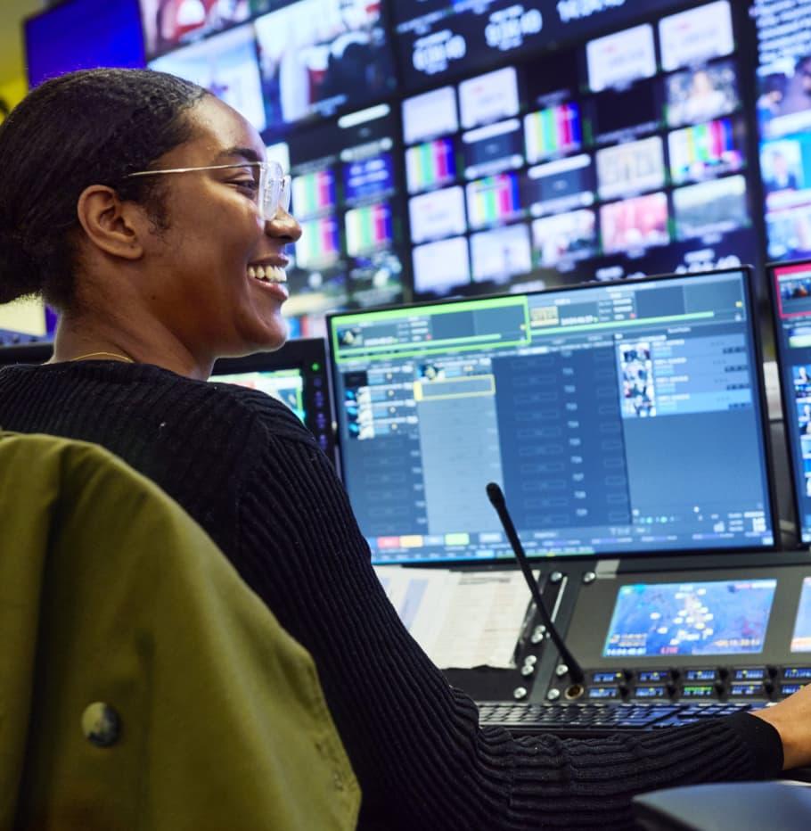 black woman in glasses smiling in front of multiple tv production screens