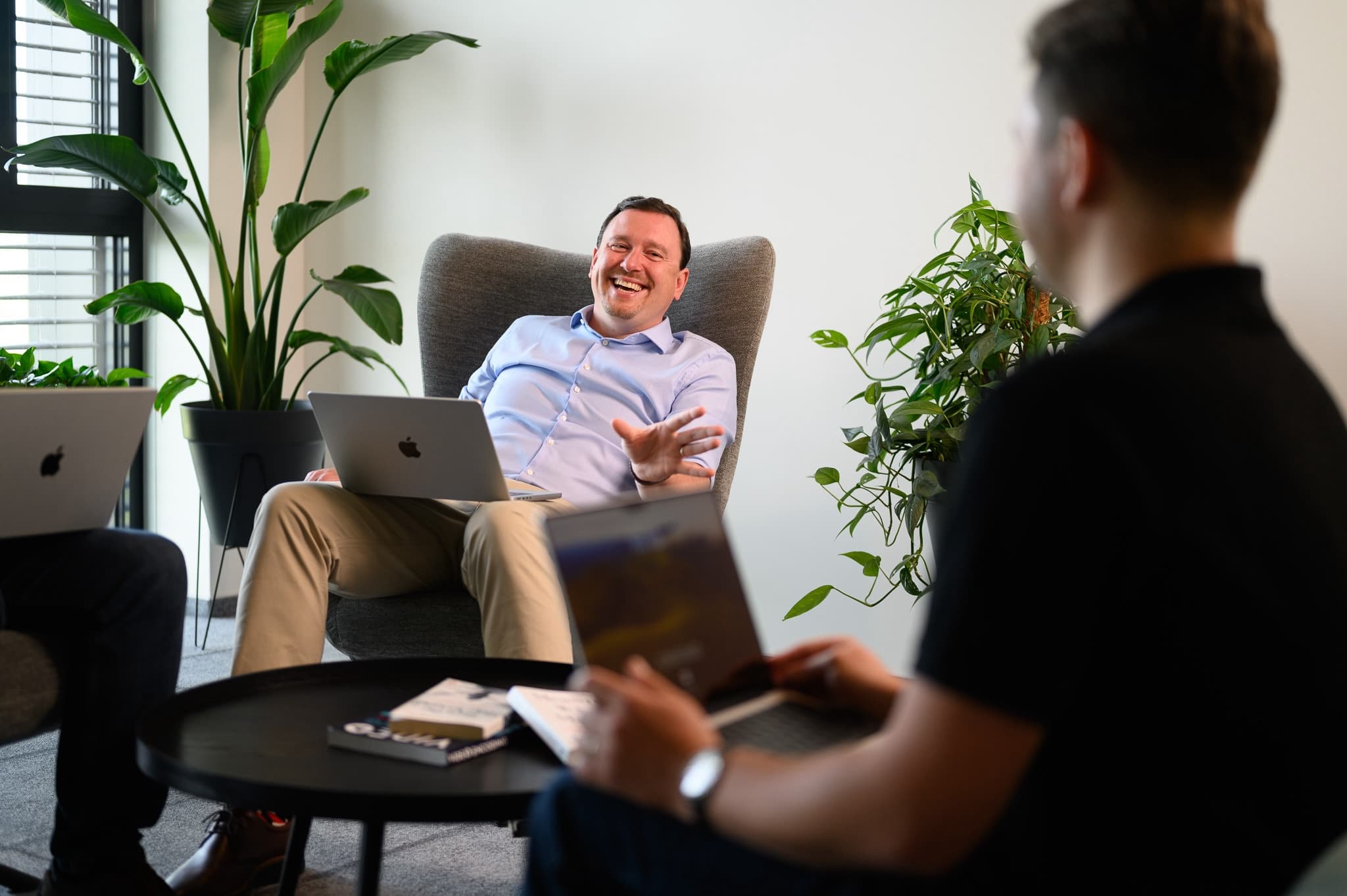 Man smiling and gesturing while seated with a laptop in a relaxed office lounge.