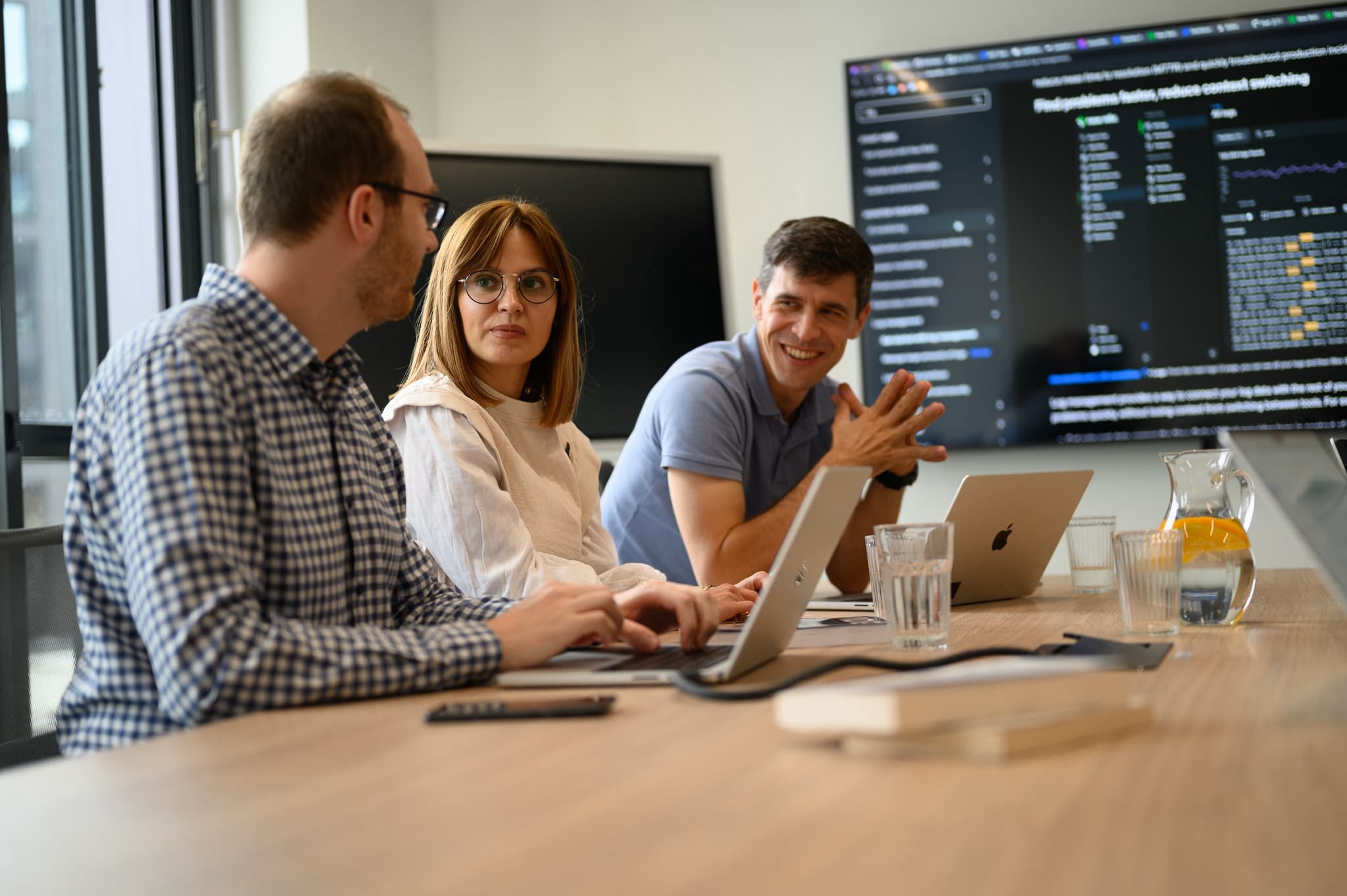 Three colleagues in a meeting room with laptops, discussing content on a large wall screen.