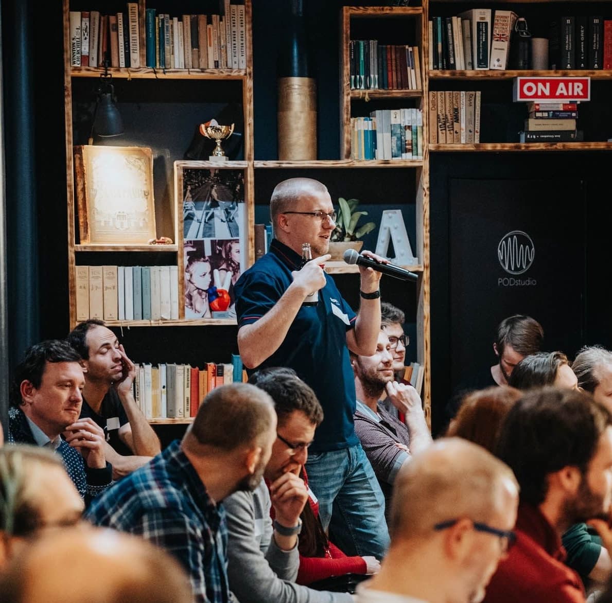 Man in a library-themed room stands and speaks into a microphone during a seated group event.