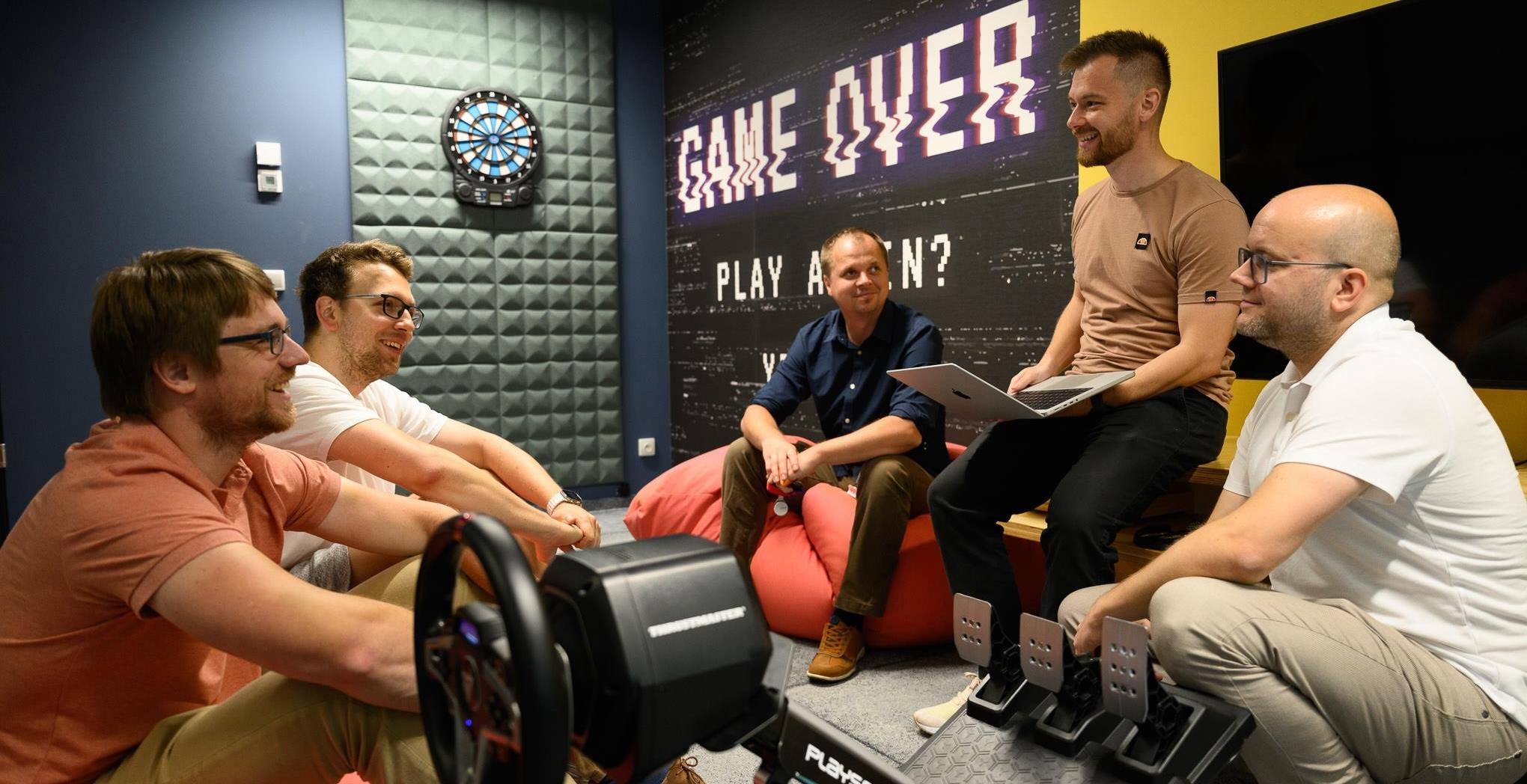 Five colleagues in a casual meeting room with a “Game Over” wall sign, chatting on bean bags and one with a laptop.