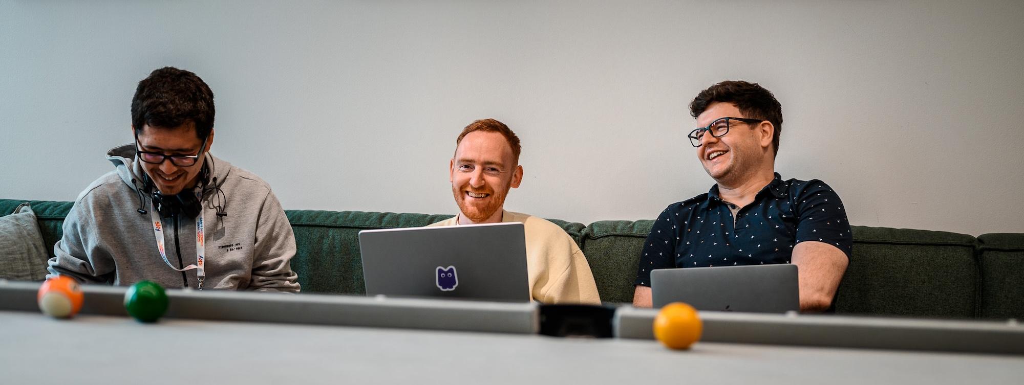 Three men on a green couch with laptops, smiling, with pool table balls in the foreground.
