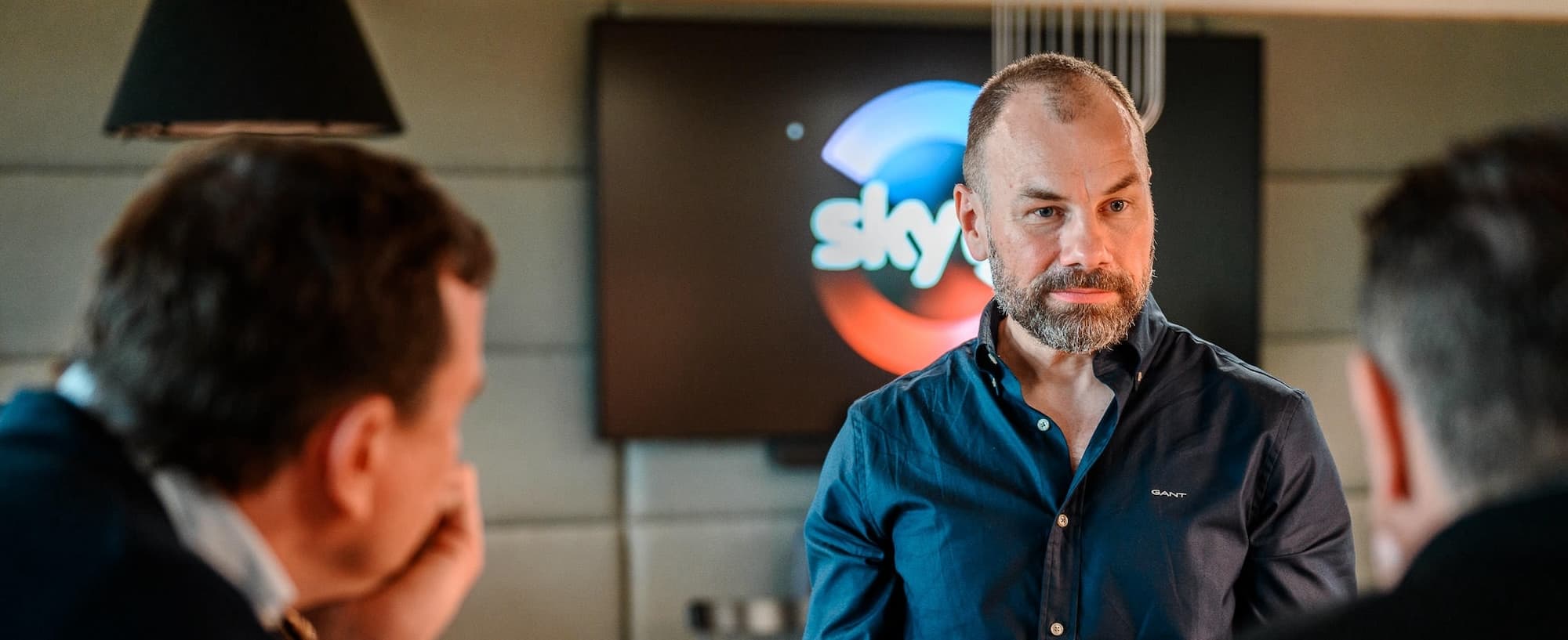 Man in a dark shirt speaking during a meeting, with a Sky logo screen behind him.