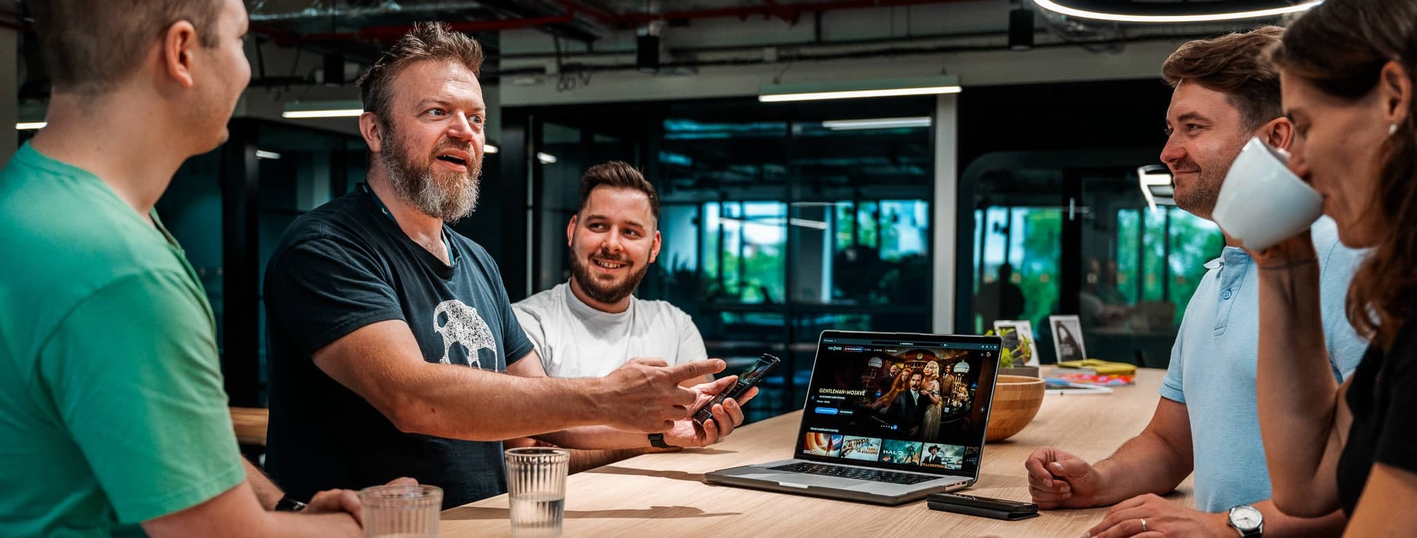 Group of five coworkers in casual discussion around a long table, one showing content on a laptop.