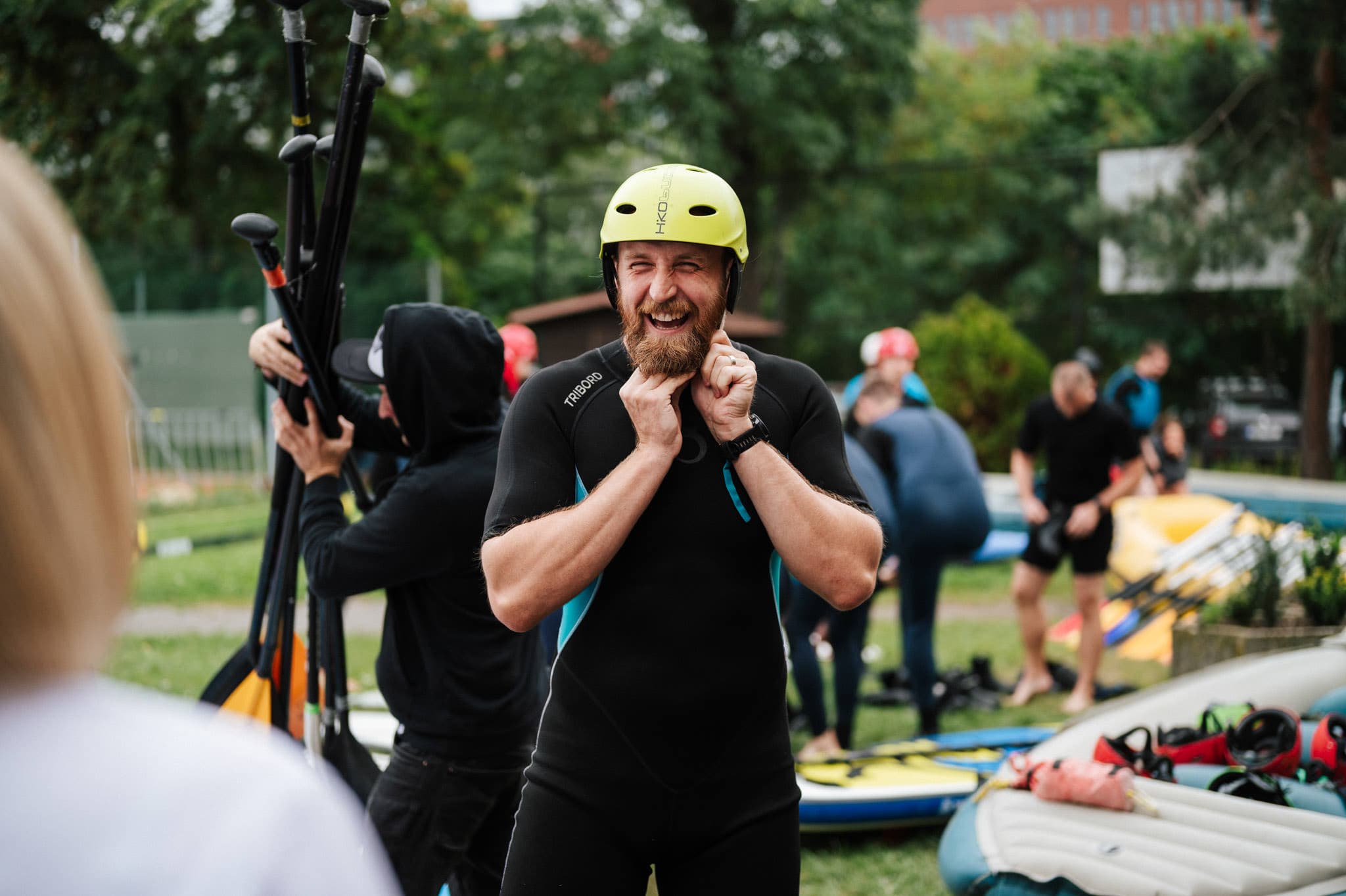 Smiling man in a wetsuit fastening a helmet, surrounded by people preparing paddleboards outdoors.