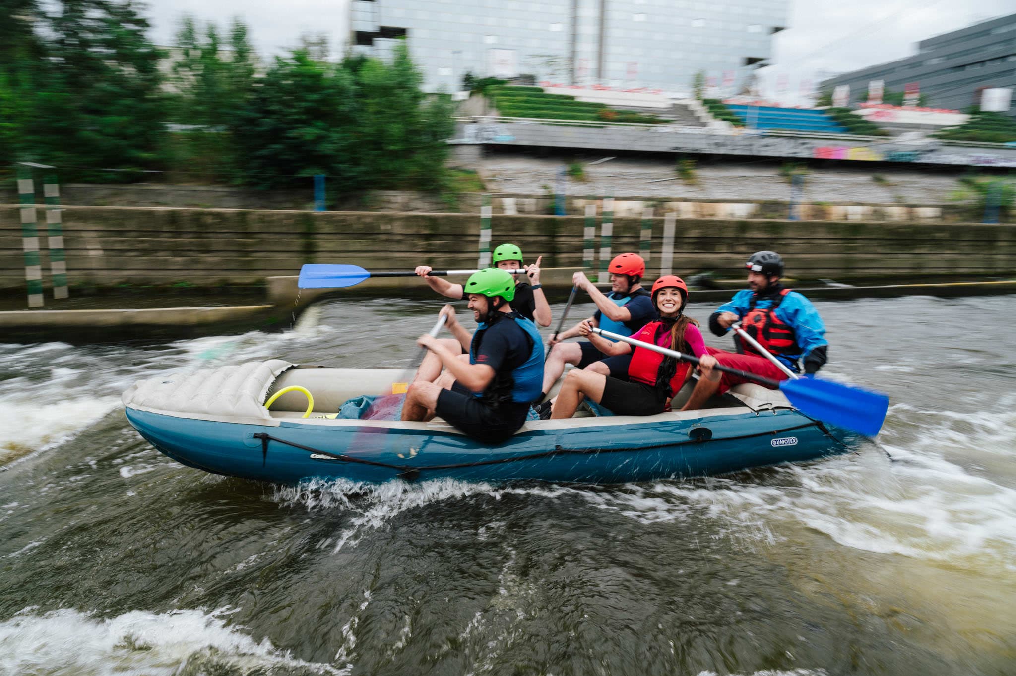 Six people whitewater rafting, all in helmets and life jackets, paddling together with big smiles in a fast-moving river.