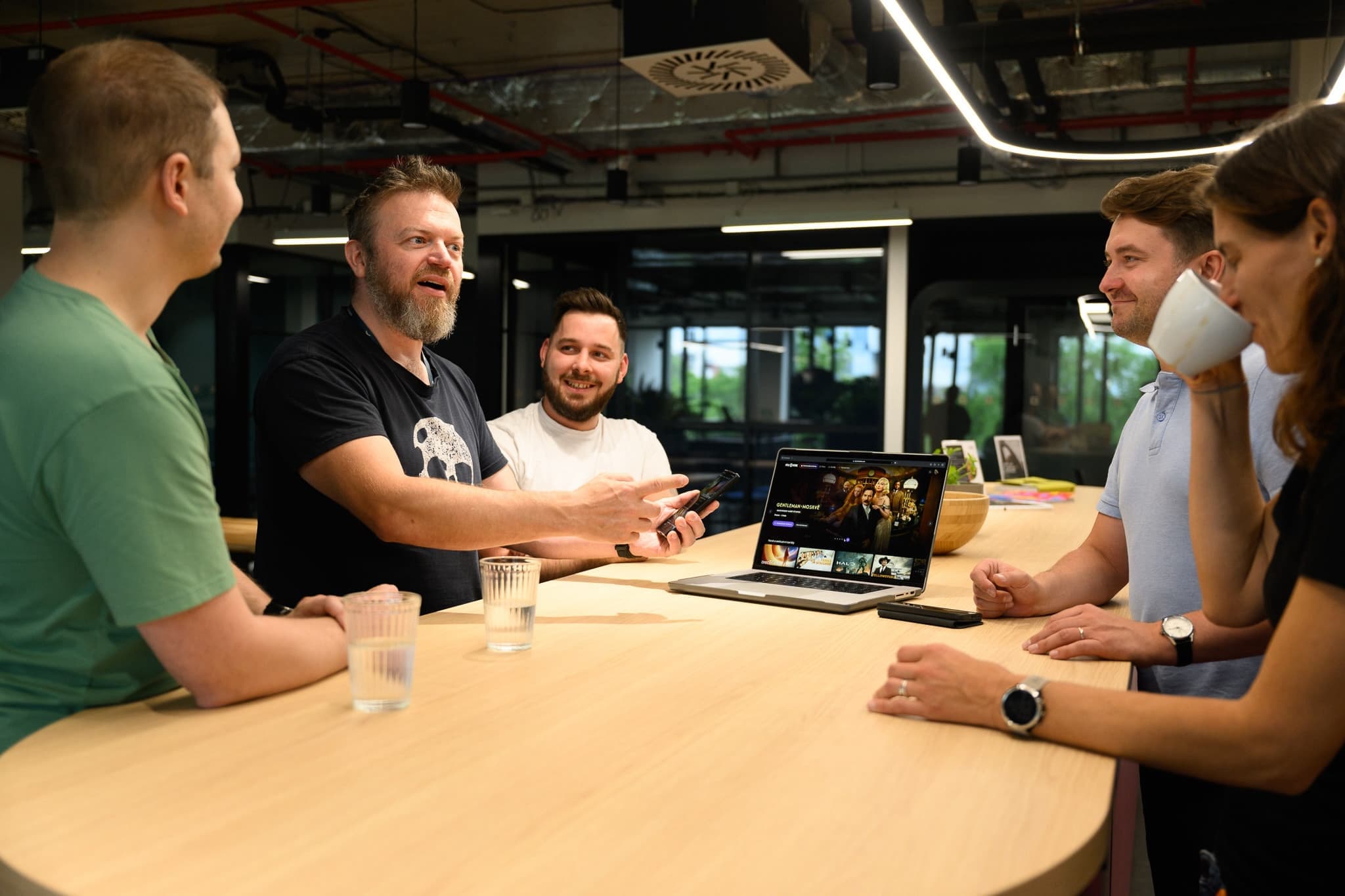 Group of five coworkers in casual discussion around a long table, one showing content on a laptop.