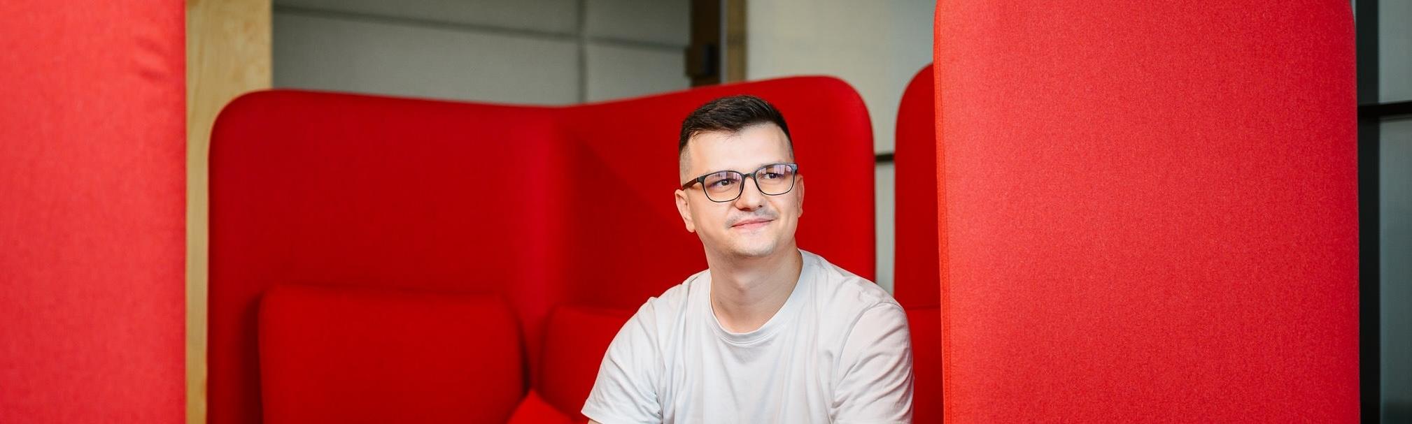 Man in a white t-shirt sitting in a modern booth with bright red cushioned walls, looking off to the side and smiling.