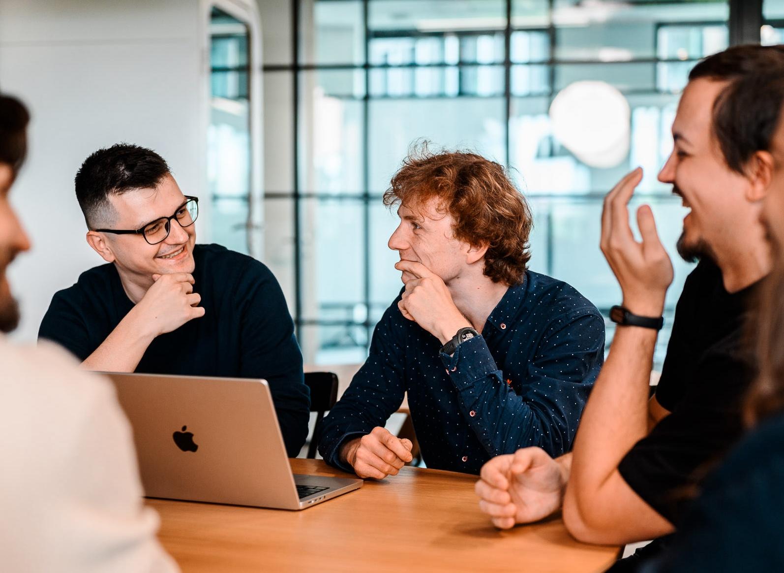Group of colleagues laughing and chatting around a table in a bright modern office.