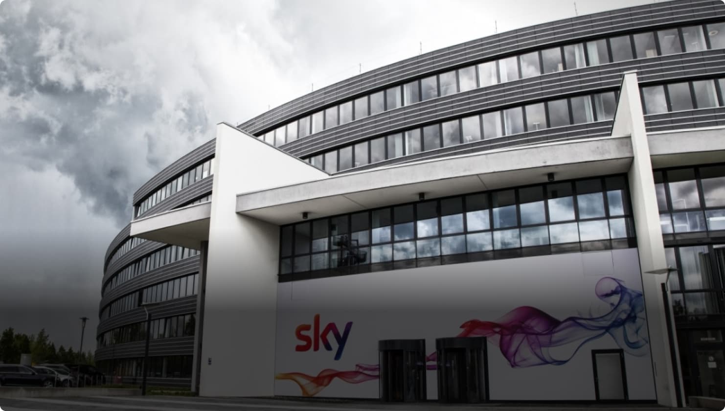 Außenansicht eines modernen Sky-Bürogebäudes mit Glasfenstern und einer Sky-Logo-Wandtapete unter einem bewölkten Himmel.