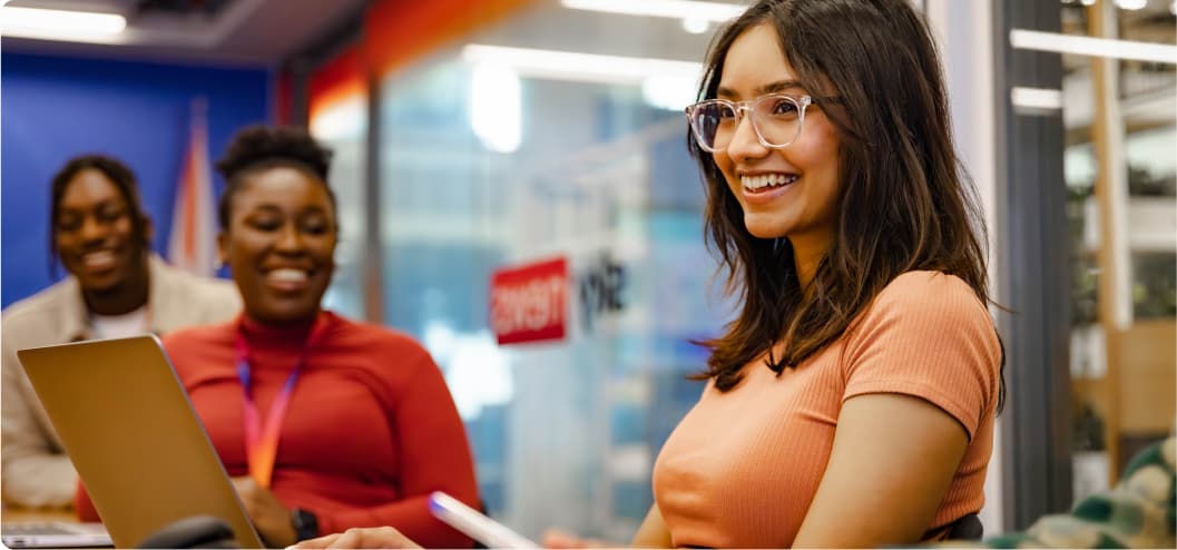 Young woman in glasses smiling while working on a laptop, with two colleagues in the background chatting in a modern office setting.