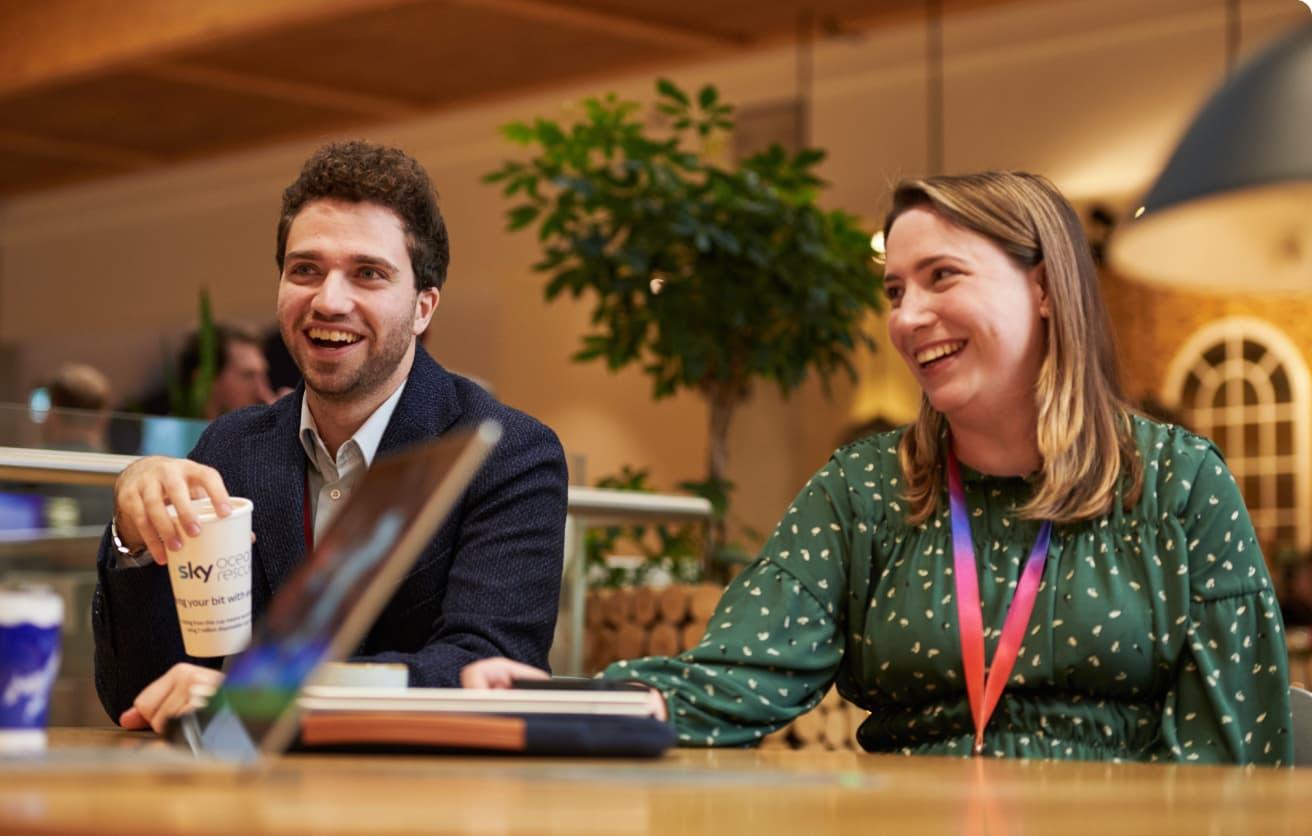 Man and woman laughing during a conversation at a wooden table in an open office café area.