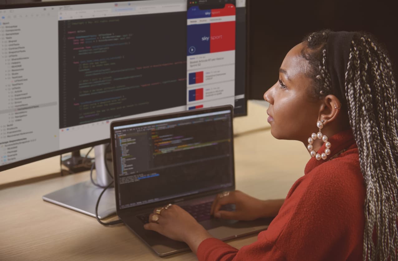 Woman with braided hair working on a coding project at her desk, with lines of code and a Sky Sports app preview visible on dual monitors.