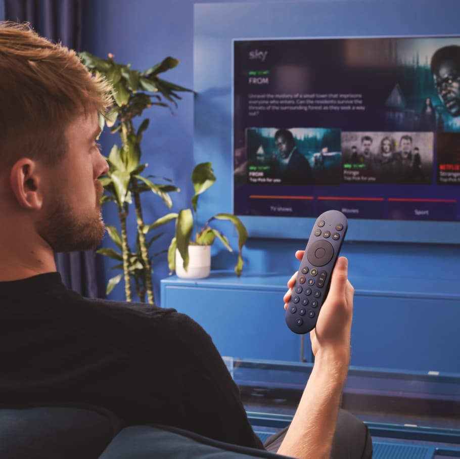 Man sitting on a couch holding a remote, facing a TV screen displaying Sky content in a cozy, blue-toned living room.
