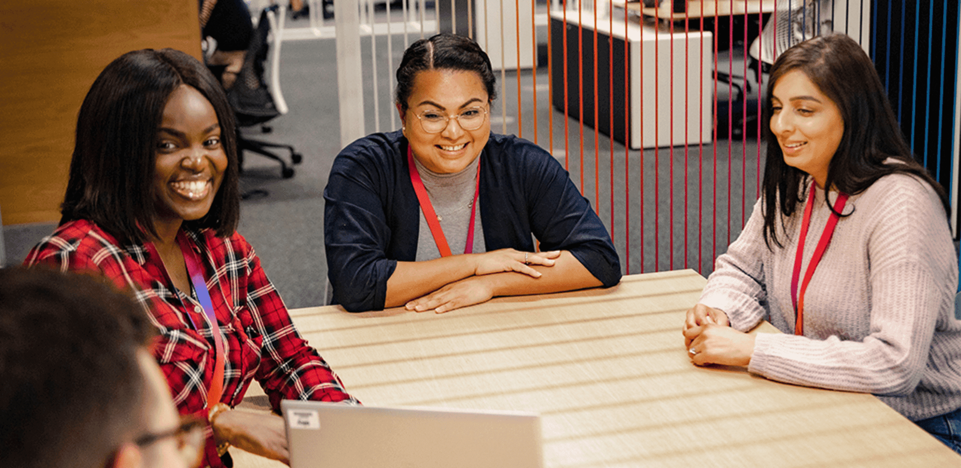 Three women seated at a table in an office, smiling and chatting during a relaxed meeting.