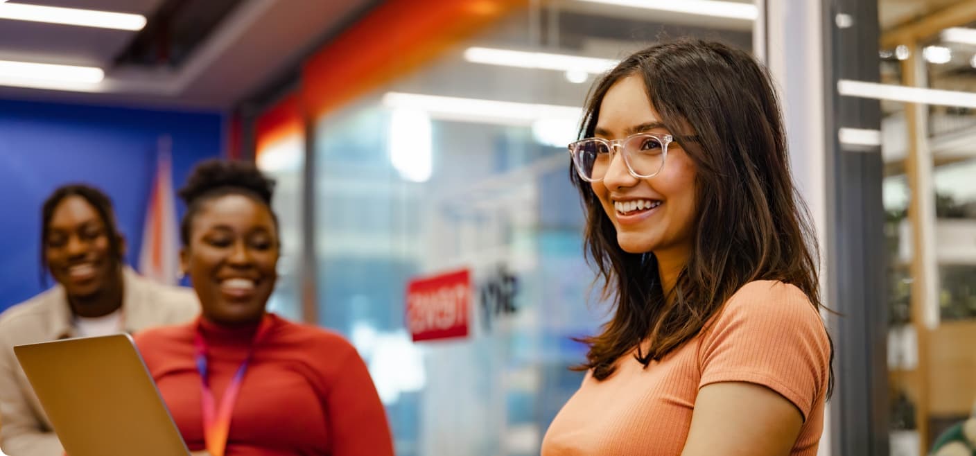 Young woman in glasses smiling while working on a laptop, with two colleagues in the background chatting in a modern office setting.