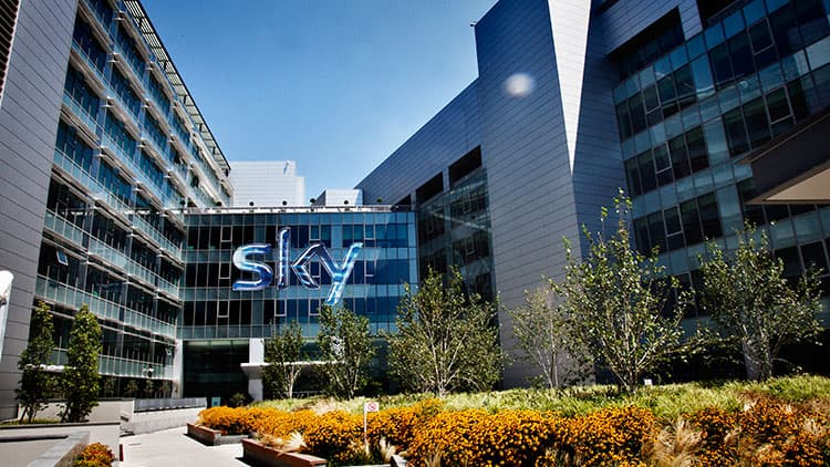 Exterior of a Sky office building with large glass windows, landscaped gardens, and a prominent illuminated Sky logo.