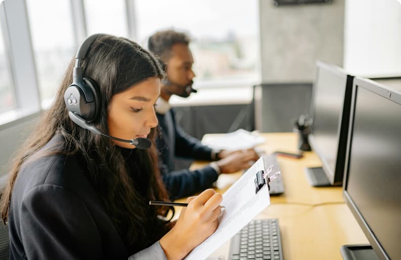 Frau mit Headset in einem Callcenter, die an ihrem Schreibtisch Notizen macht, neben ihr ein männlicher Kollege, der an einem PC arbeitet.