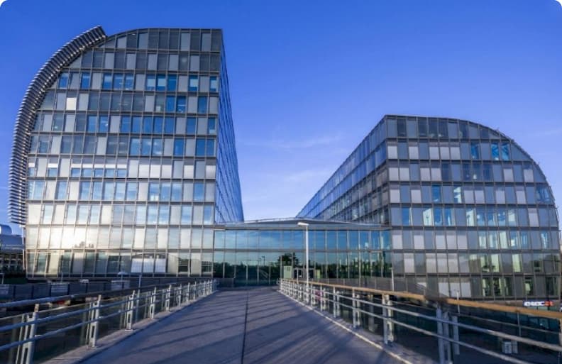 Modernes Bürogebäude aus Glas mit geschwungenen Fassaden und einem Gang, der zum Haupteingang führt, vor einem klaren blauen Himmel.