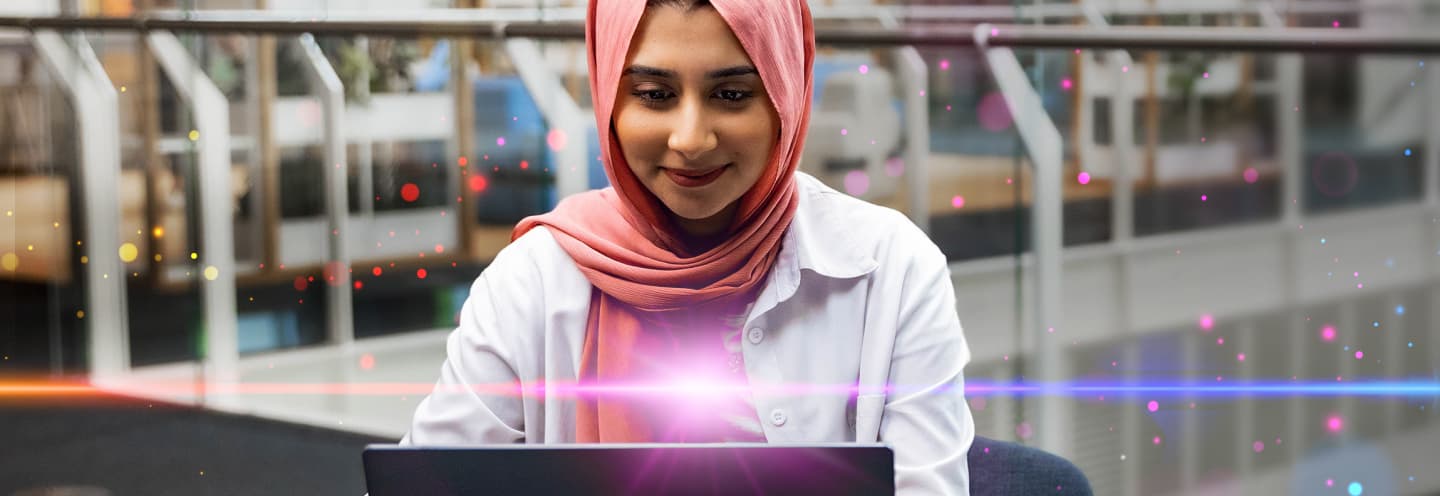 Eine Frau mit einem rosa Kopftuch arbeitet an einem Laptop, umgeben von bunten Leuchteffekten.
