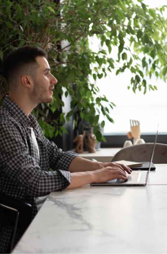 Mid shot image of a white male in a plaid shirt sitting in a bright open office with a laptop. There is a bright green tree behind him with a window