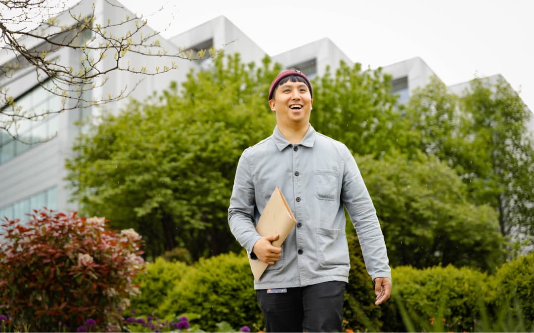 Image of a smiling man walking outside in a green area. He is wearing a blue button up top and a red hat.