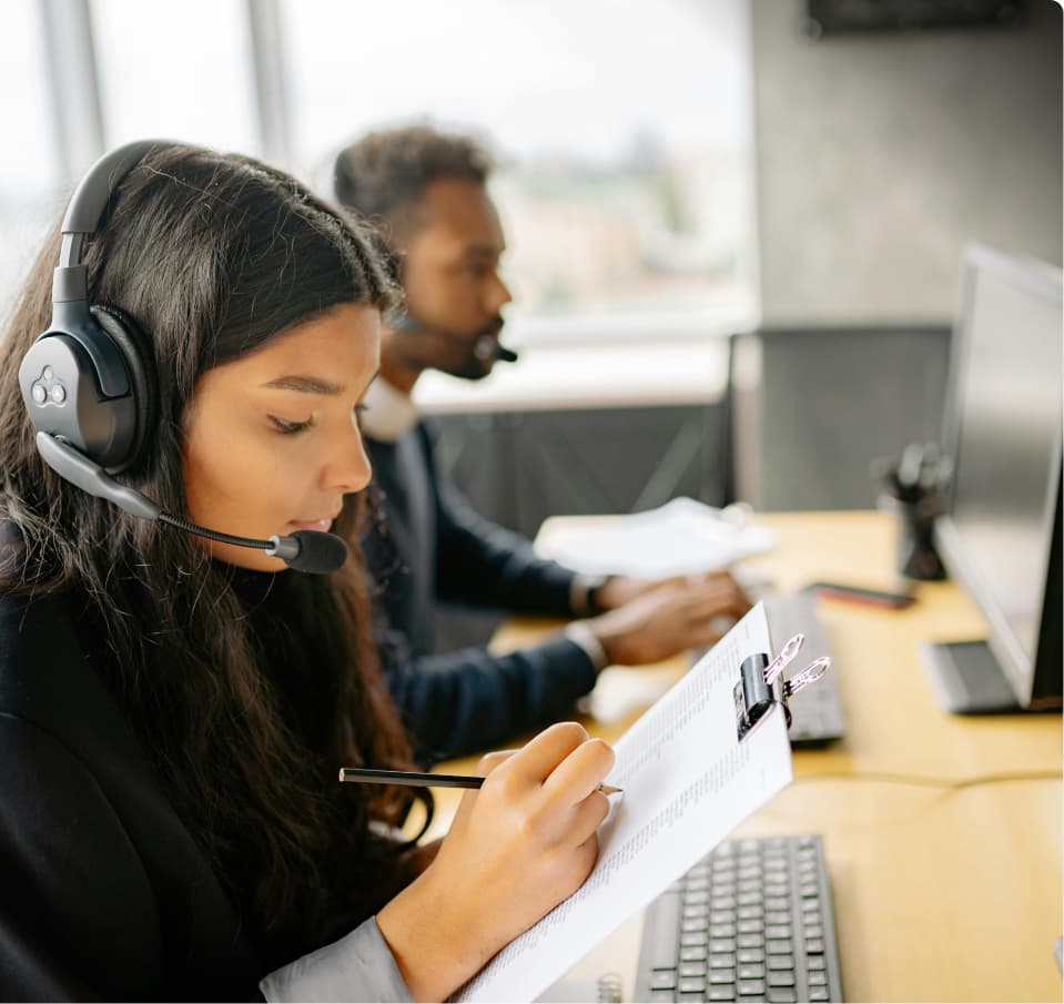 Woman in a call centre headset writing notes on a clipboard at her desk, with a male colleague beside her working on a desktop computer.