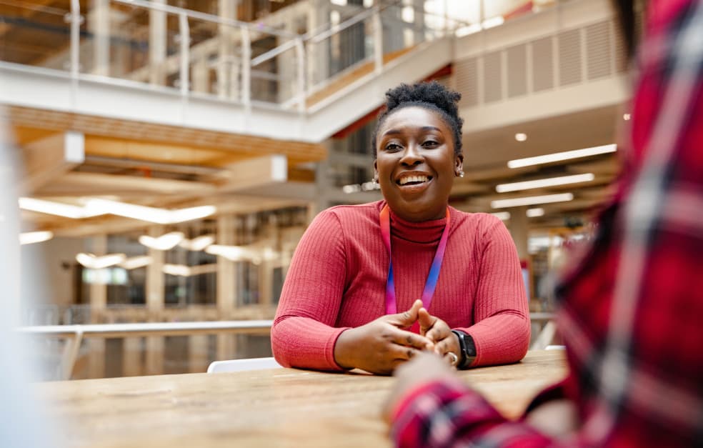 Mid shot focused image of a black women sat in an office. She is wearing a pink roll-neck and lanyard, and smiling.