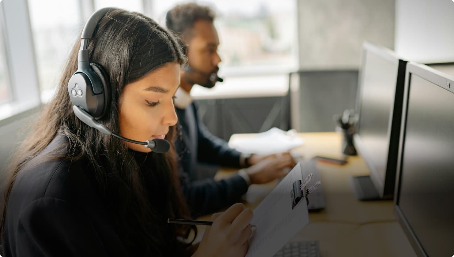 Frau mit Headset in einem Callcenter, die an ihrem Schreibtisch Notizen macht, neben ihr ein männlicher Kollege, der an einem PC arbeitet.
