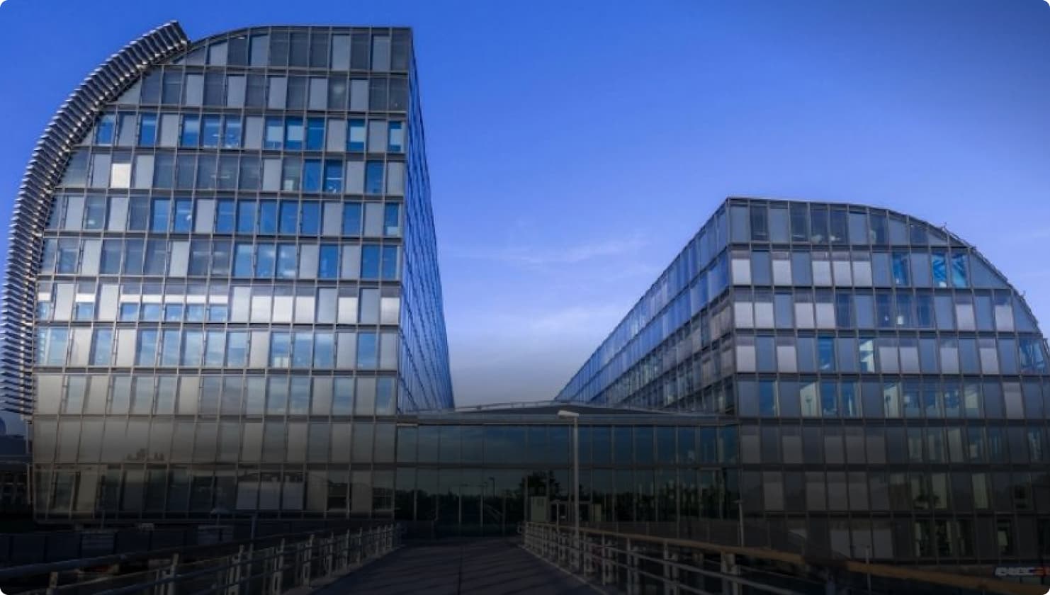 Modernes Bürogebäude aus Glas mit geschwungenen Fassaden und einem Gang, der zum Haupteingang führt, vor einem klaren blauen Himmel.
