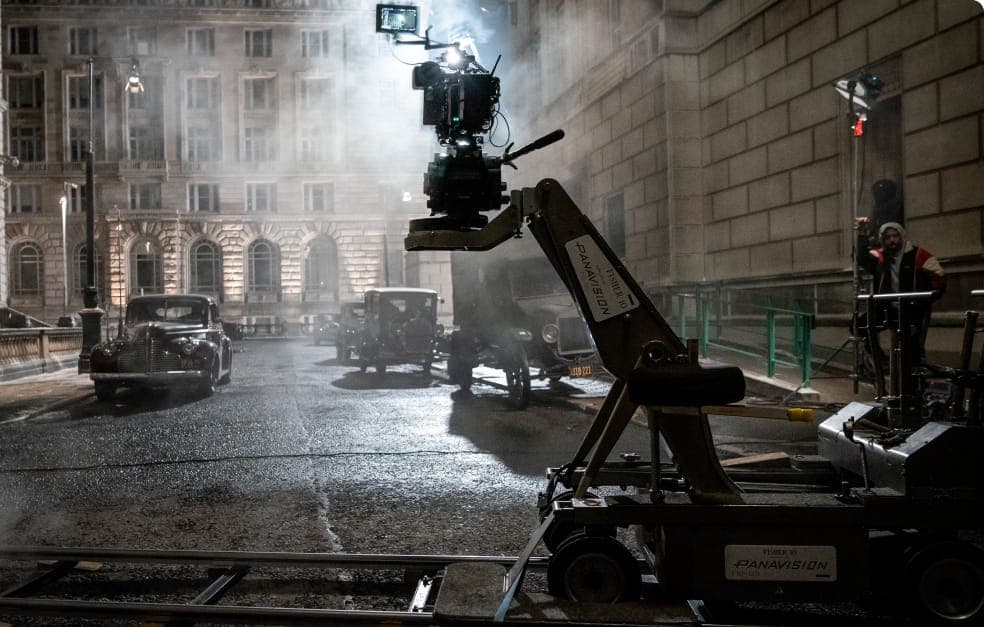 Film production setup on a city street at night, with a large camera crane, vintage cars, and atmospheric lighting with rising smoke.