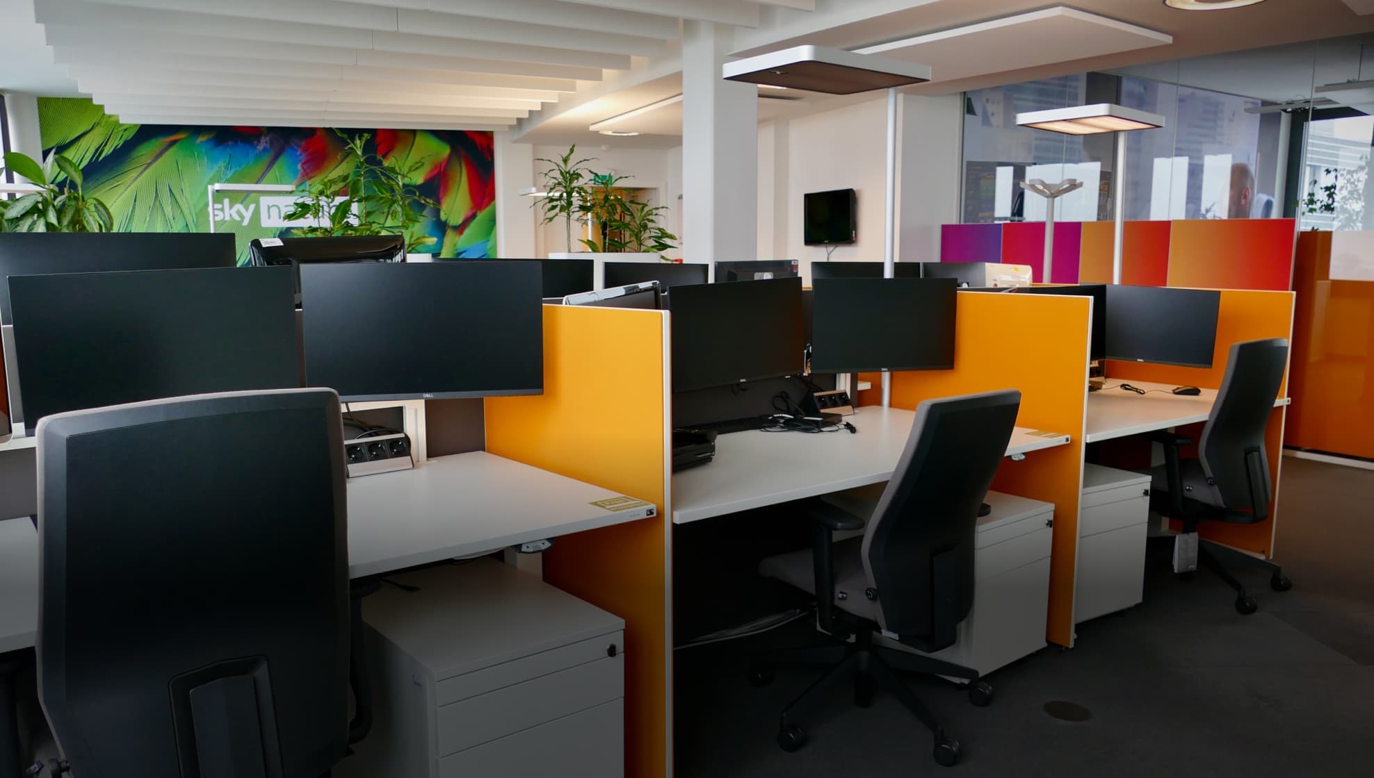 Modern open-plan office with orange partitioned desks, multiple computer monitors, and green plants under a bright ceiling.