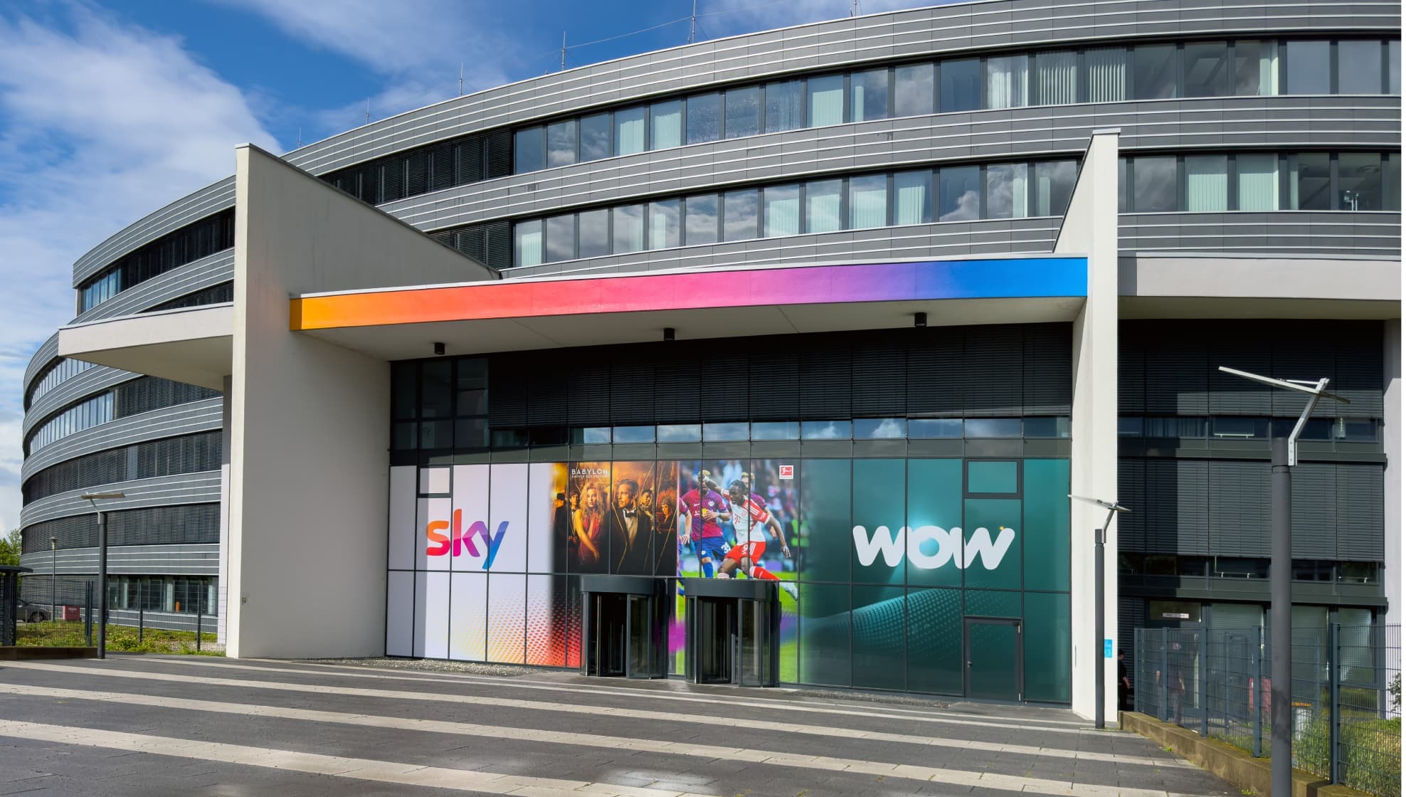 Exterior of the Sky Germany building with a curved facade, rainbow-colored trim, and branded Sky and WOW signs on the glass.