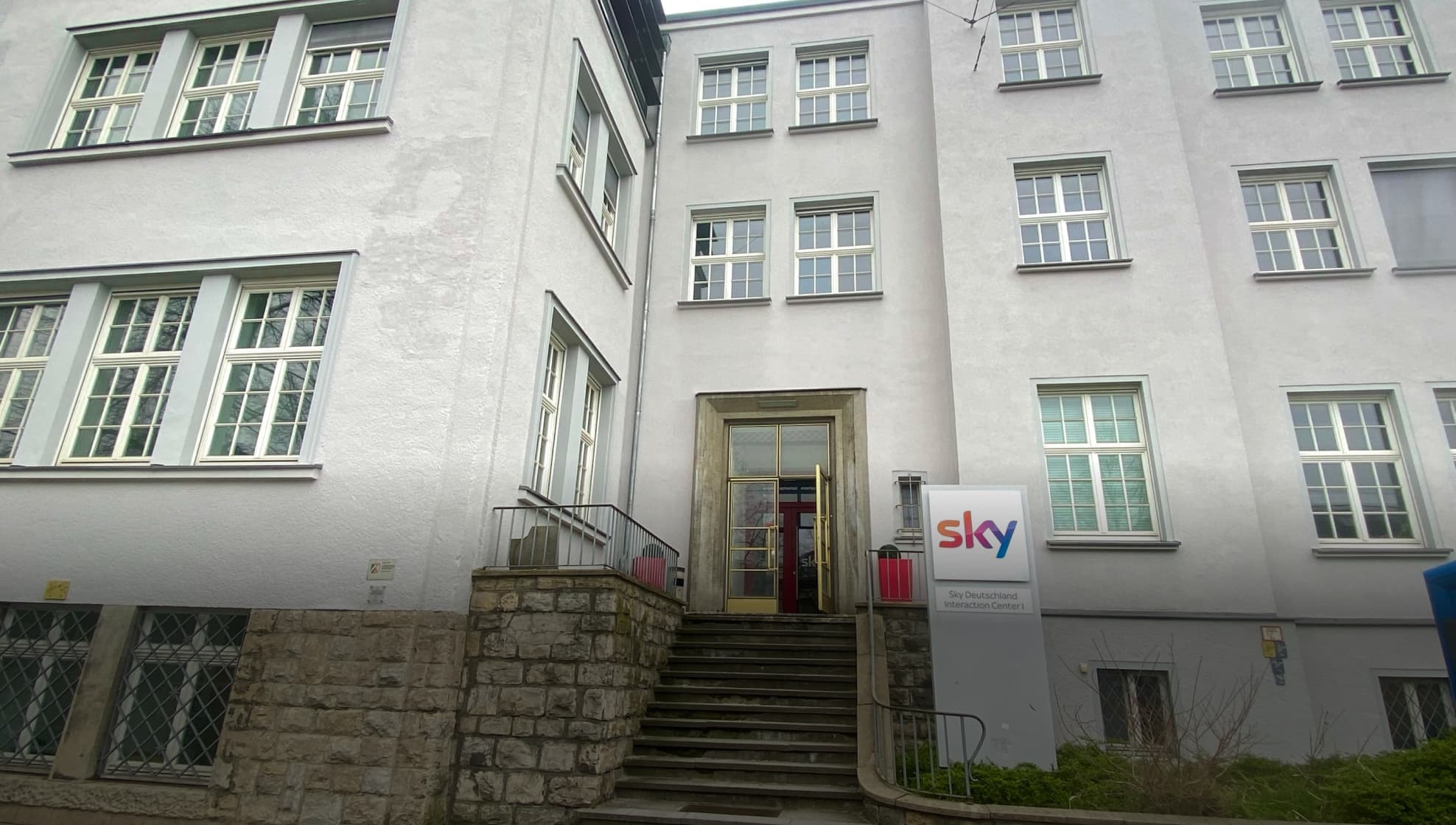 Eingang zum Sky Deutschland Interaction Center I, einem hellgrauen Bürogebäude mit Stufen und einem Sky-Schild.