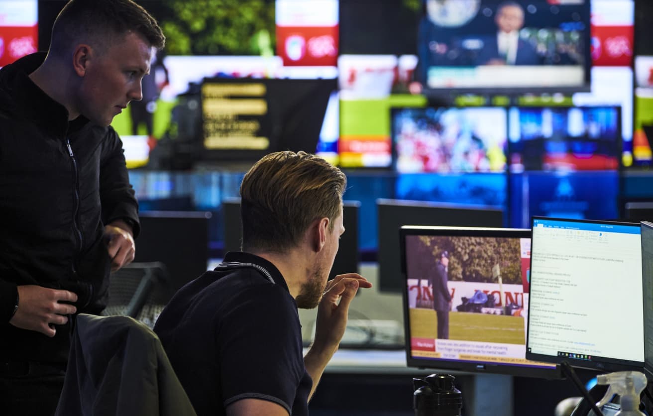 Two men in a broadcast control room focused on monitors, with multiple TV screens displaying news and sports content in the background.Two men in a broadcast control room focused on monitors, with multiple TV screens displaying sports content in the background.