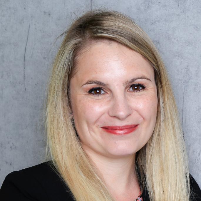 Portrait of a smiling woman with long blonde hair, wearing a black blazer, in front of a grey wall.