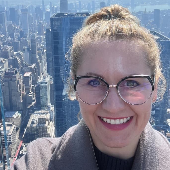 Smiling woman in glasses and a coat, with a city skyline behind her.