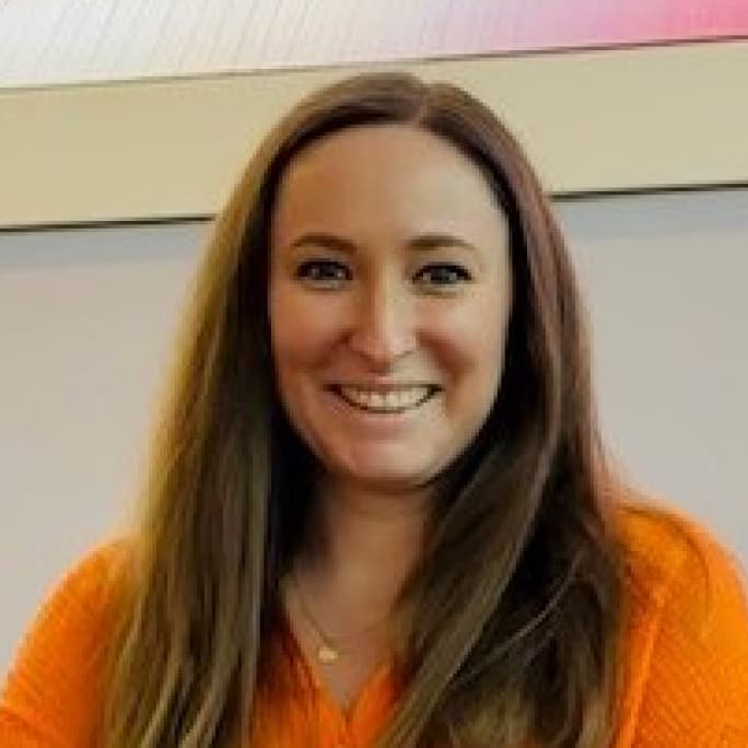 Woman smiling at the camera, wearing an orange top and long straight hair, seated indoors.