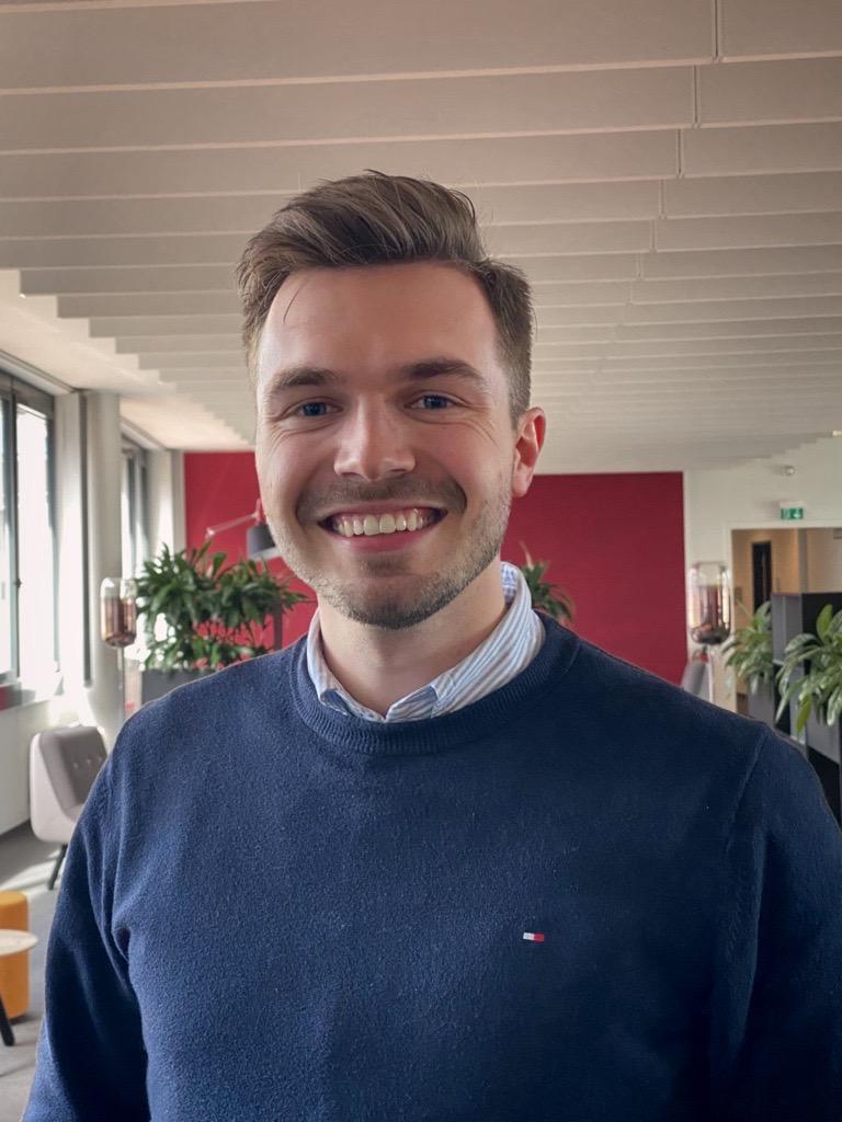Man smiling in a navy sweater, standing in a modern office space with plants and red walls.