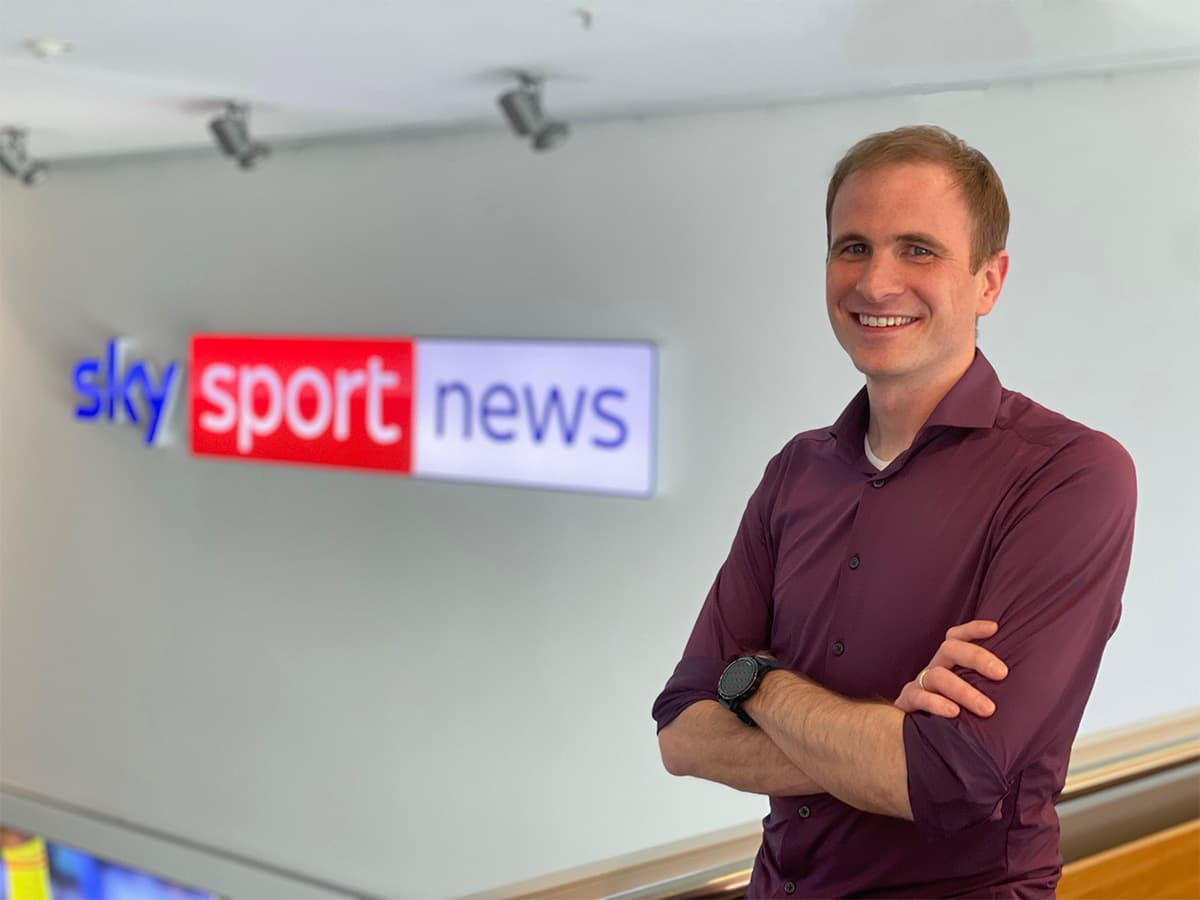 Phillip stands in a burgundy-coloured shirt, with his arms crossed. In the background is the Sky Sport News signage fixed onto the wall.