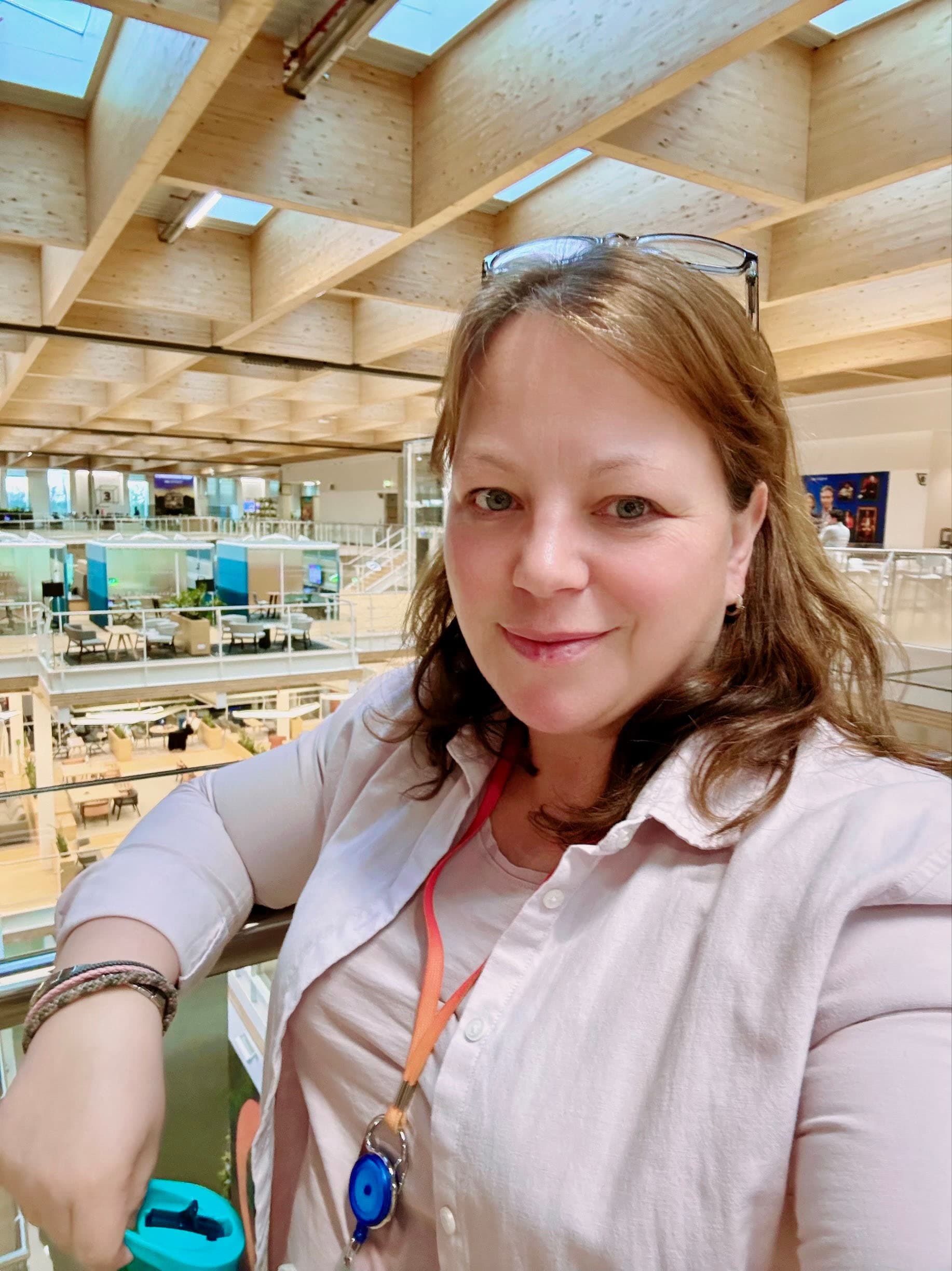 Raika is standing against a metal guard-rail, overlooking the Sky Central campus in Osterley, London. Her right arm is resting on the rail, as colleagues across three floors in the background enjoy meetings in the many breakout spaces in the distance.