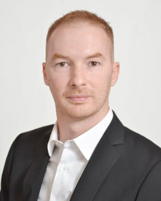 Professionelles Foto eines Mannes in einem schwarzen Blazer und weißem Hemd, neutraler Hintergrund.