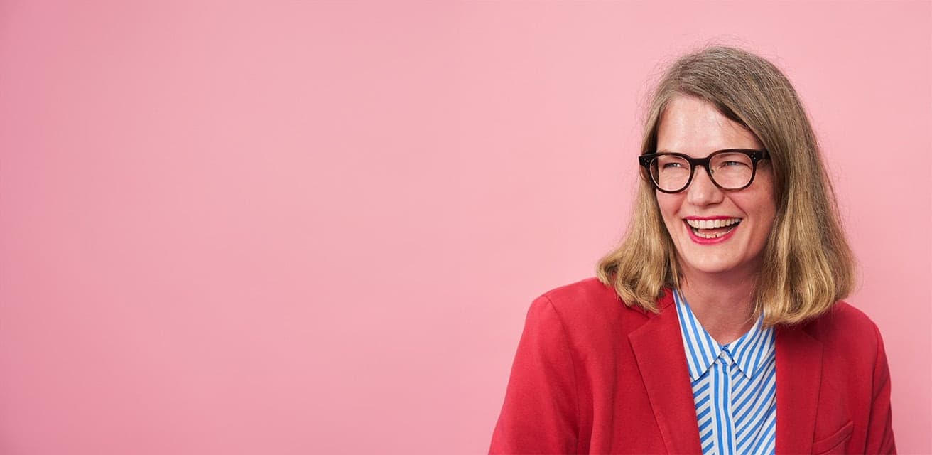 Smiling woman wearing glasses and a red blazer over a blue-striped shirt, posed in front of a solid pink background.