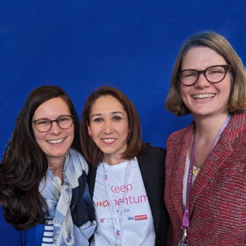Three women smiling in front of a blue backdrop, wearing Sky event lanyards.