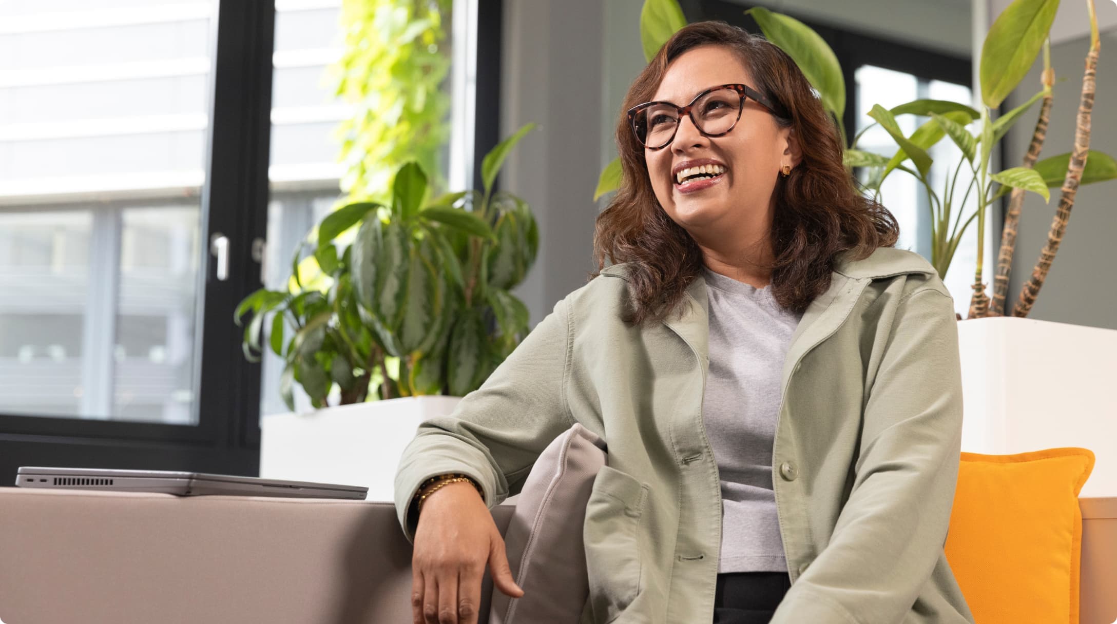 Woman smiling while seated in a lounge area with plants and natural light, laptop resting on the couch beside her.