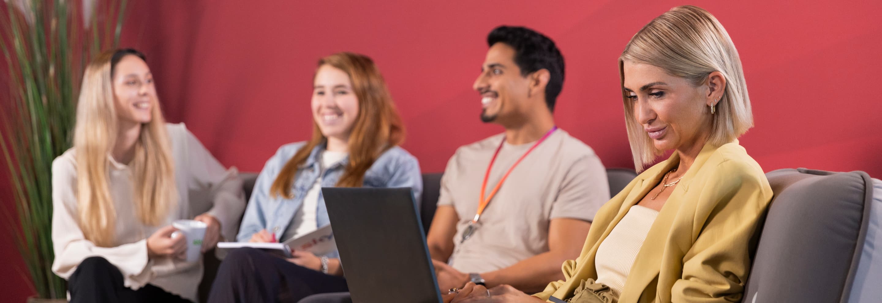 Four colleagues seated on a couch with a red wall behind, one woman focused on her laptop while others smile and talk.