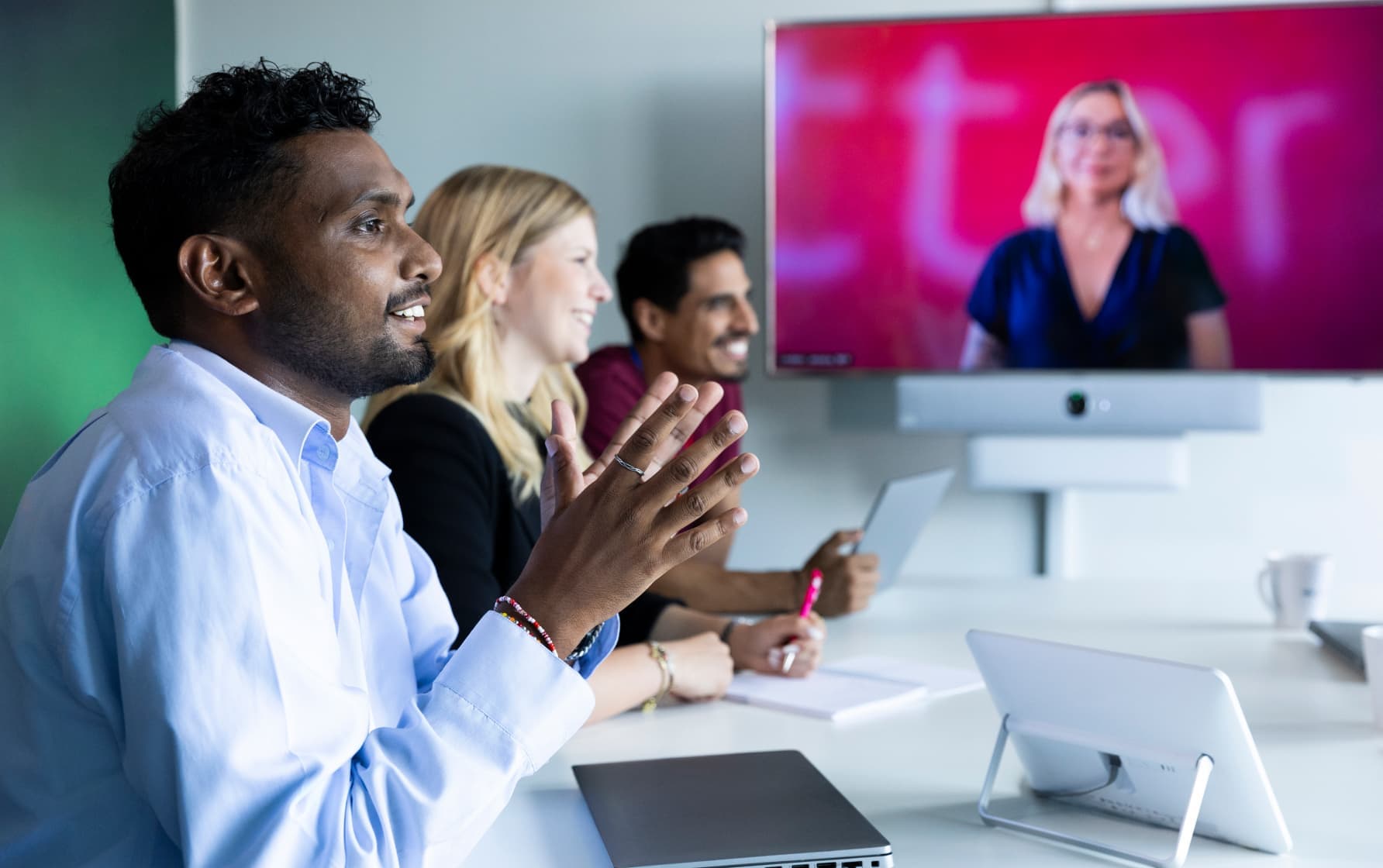 Colleagues seated in a conference room attending a hybrid meeting, with a woman visible on a video call screen in front of them.