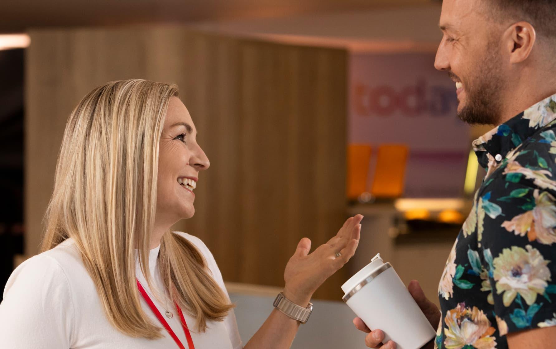 Two colleagues smiling and chatting indoors, one holding a reusable cup, with a blurred office background.