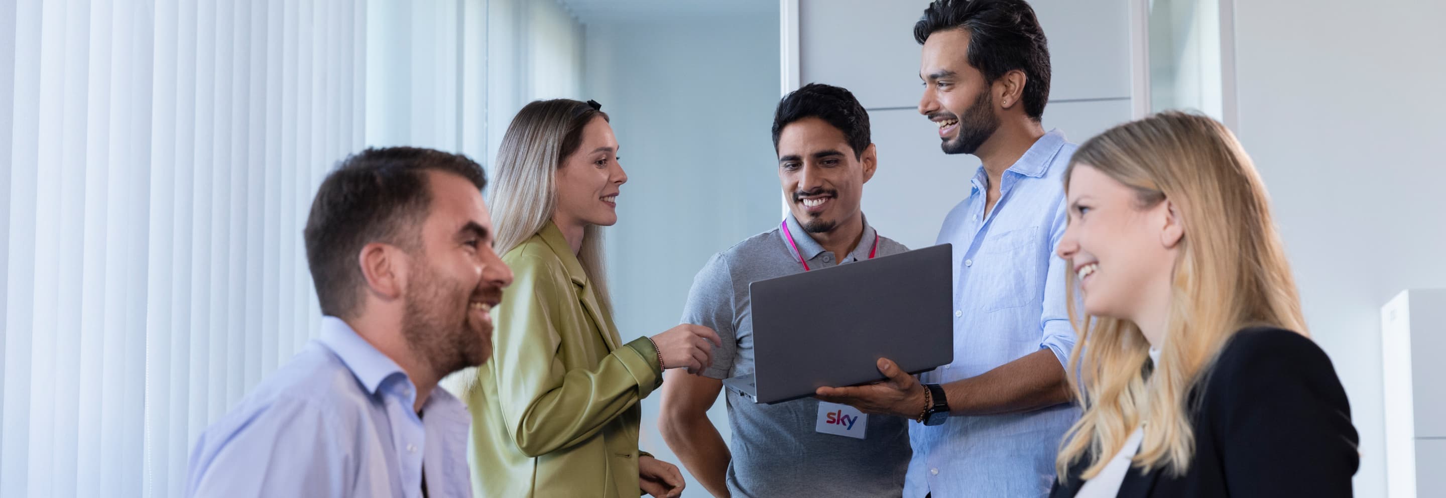 Group of five smiling colleagues chatting around a laptop in a bright office, with name badges visible and natural light coming through vertical blinds.