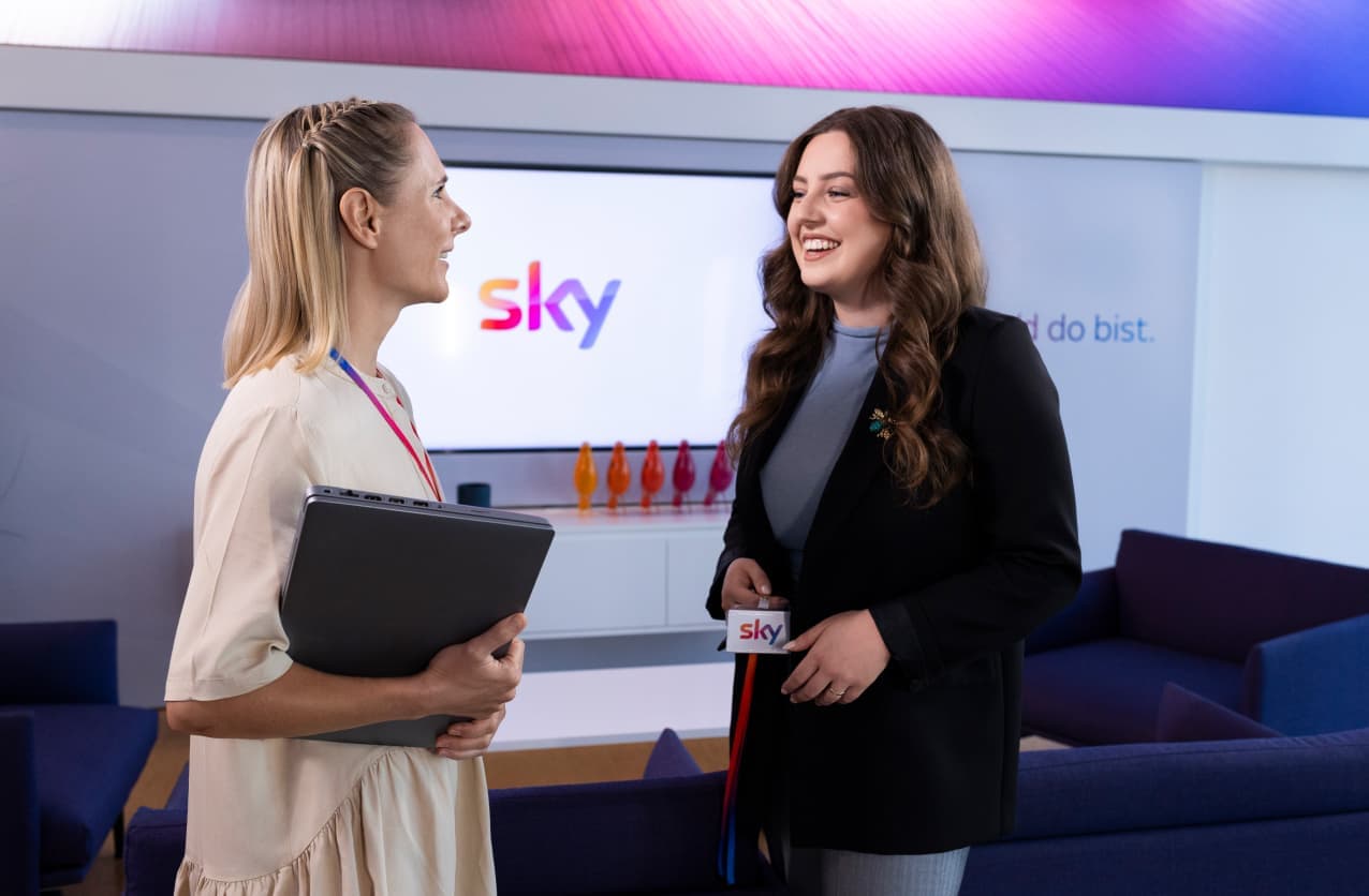 Two women standing and smiling in a brightly lit office lounge, one holding a laptop and the other a Sky staff badge.